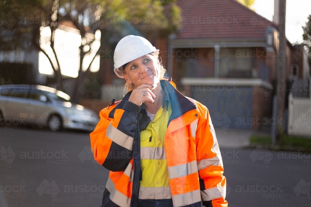 Woman road worker looking up with a white helmet and orange jacket with silver reflector on it - Australian Stock Image