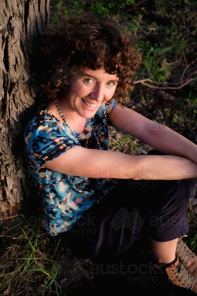 Woman sitting against trunk of tree in golden light - Australian Stock Image
