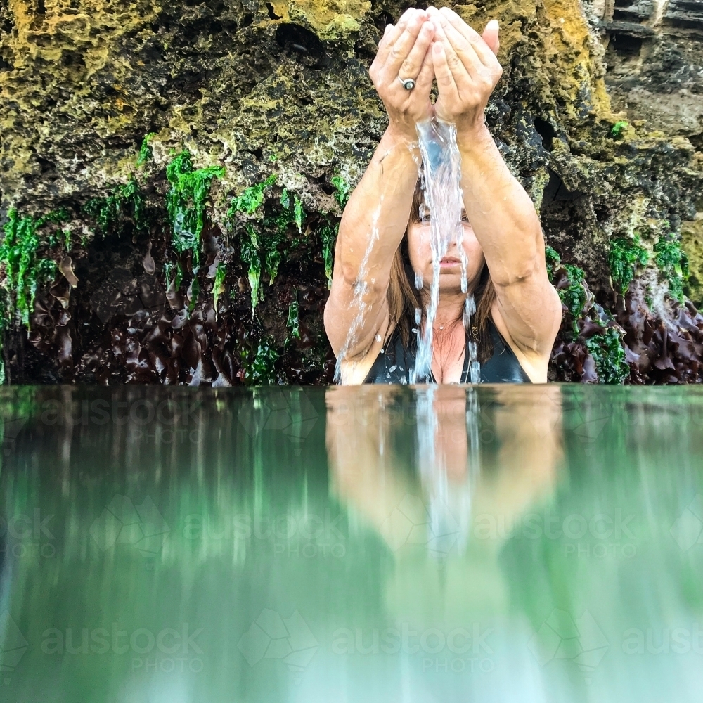 Woman sitting in glassy ocean and cupping water with coastal limestone rocks in background - Australian Stock Image