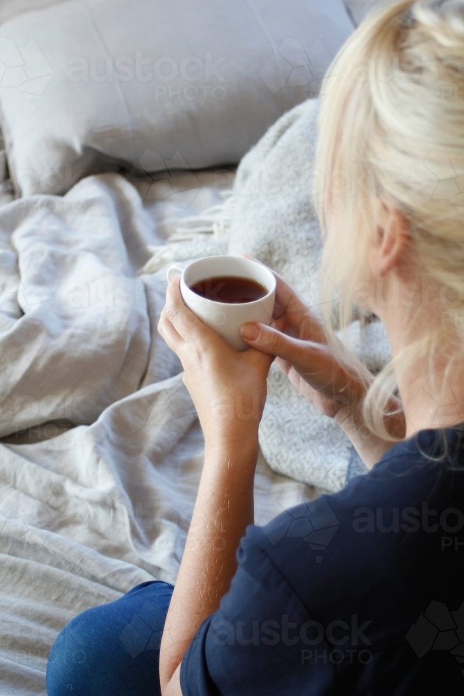 Woman sitting on bed holding cup of tea or coffee - Australian Stock Image