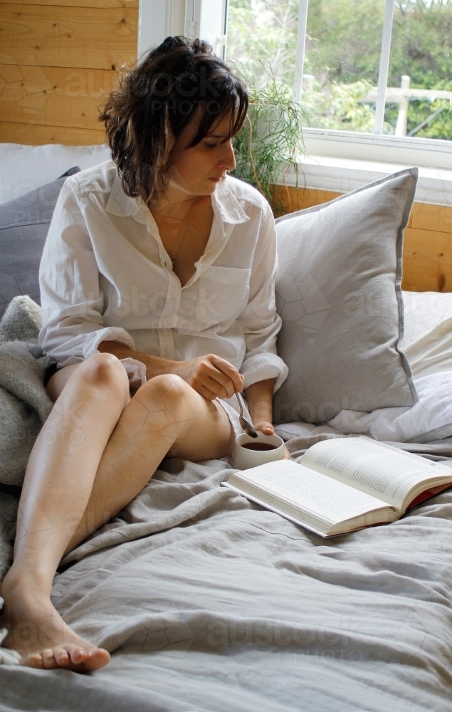 Woman sitting on bed reading book in morning - Australian Stock Image