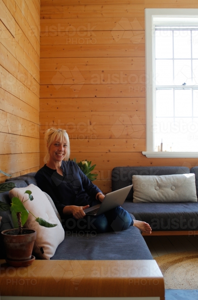 Woman sitting on couch working on laptop with timber wall background - Australian Stock Image
