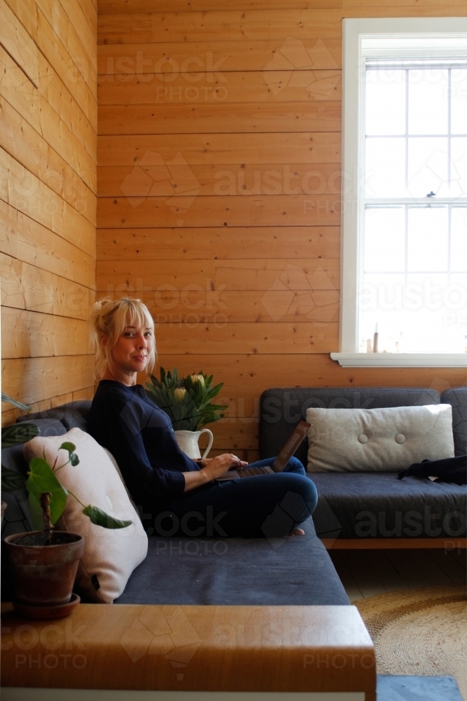 Woman sitting on couch working on laptop with timber wall background - Australian Stock Image