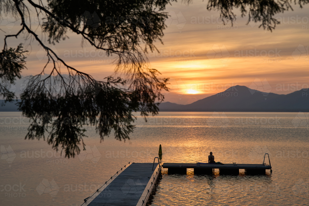 woman sitting on jetty watching sunset with tree in foreground - Australian Stock Image