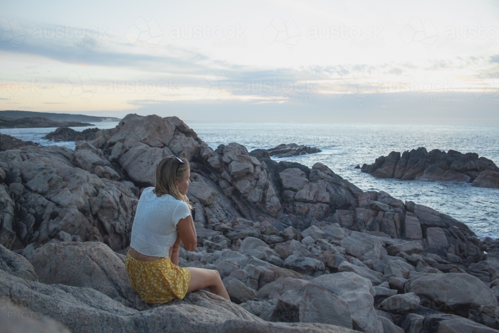 Woman sitting on rocky coastline of South West WA looking out over ocean - Australian Stock Image