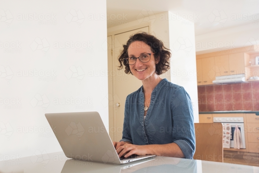 Woman smiling at camera while working on her computer at home. - Australian Stock Image