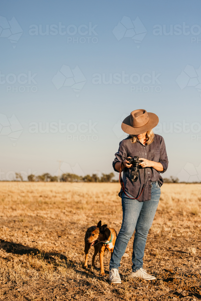 Woman standing in a dusty and dry grassy farm paddock holding a camera - Australian Stock Image