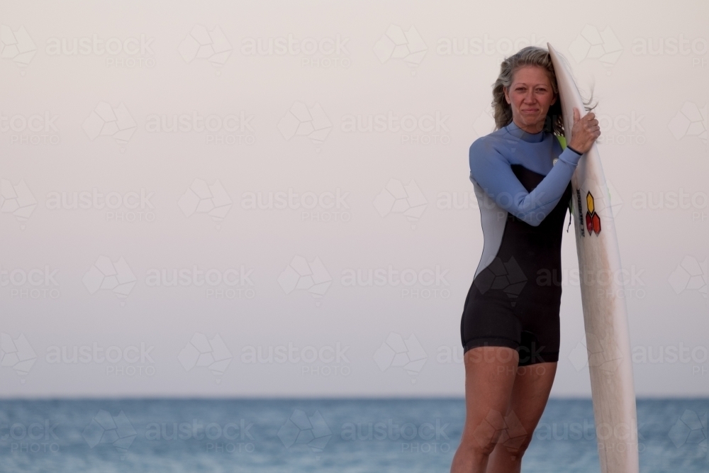 Woman Surfer standing, holding surfboard alongside body with ocean in background - Australian Stock Image