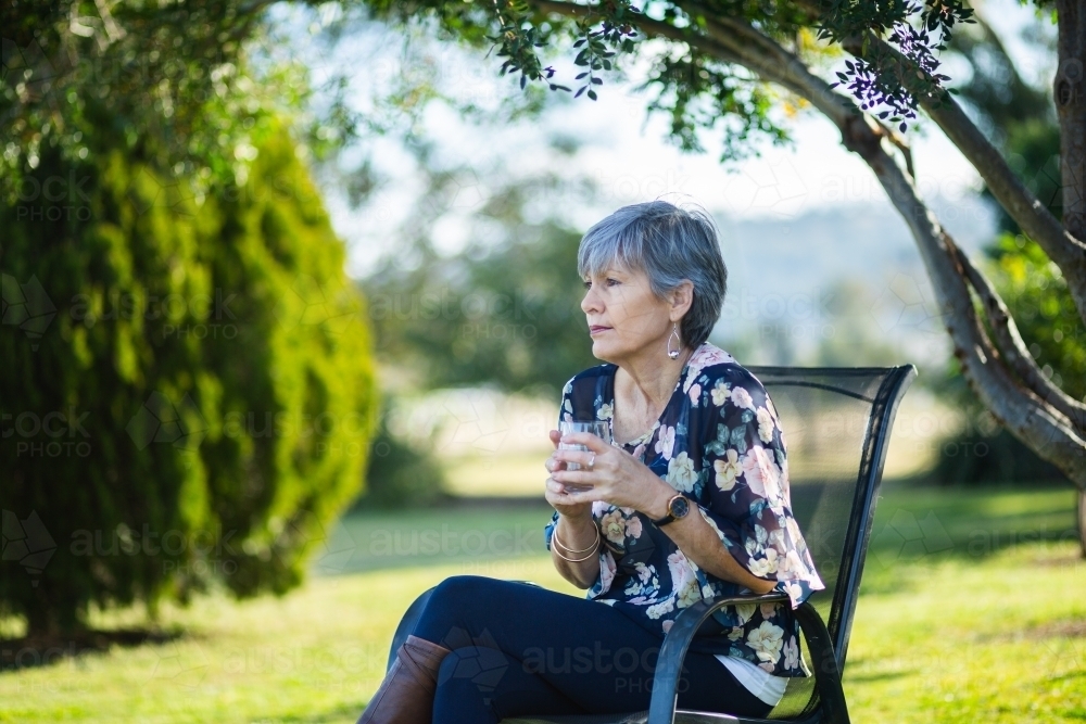 Woman with a glass of water sitting in chair under shady tree - Australian Stock Image