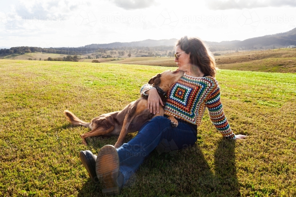 woman with arm around pet kelpie dog resting on lawn on farm playing together - Australian Stock Image