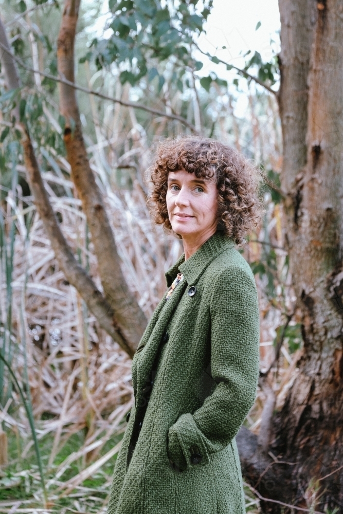 Woman with curly hair posing in front of bushland and reeds grass wearing green trench coat - Australian Stock Image