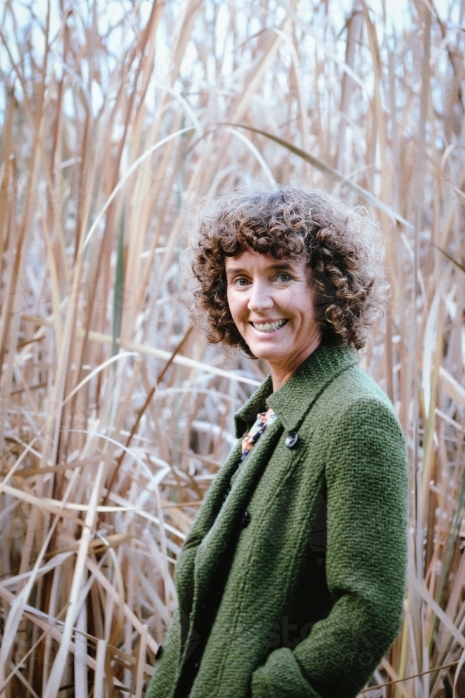 Woman with curly hair posing in front of bushland and reeds grass wearing green trench coat - Australian Stock Image