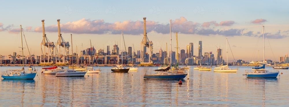 Yachts anchored on still bay in front of large gantry cranes at a cargo dock - Australian Stock Image