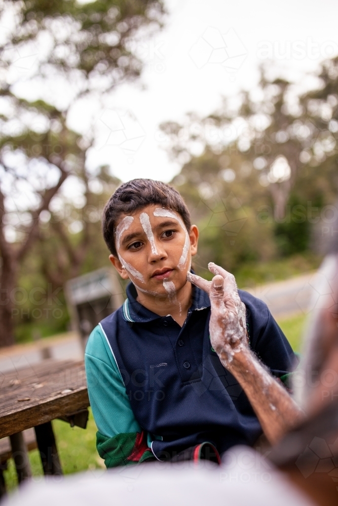 Young Aboriginal boy getting white face paint outdoors - Australian Stock Image