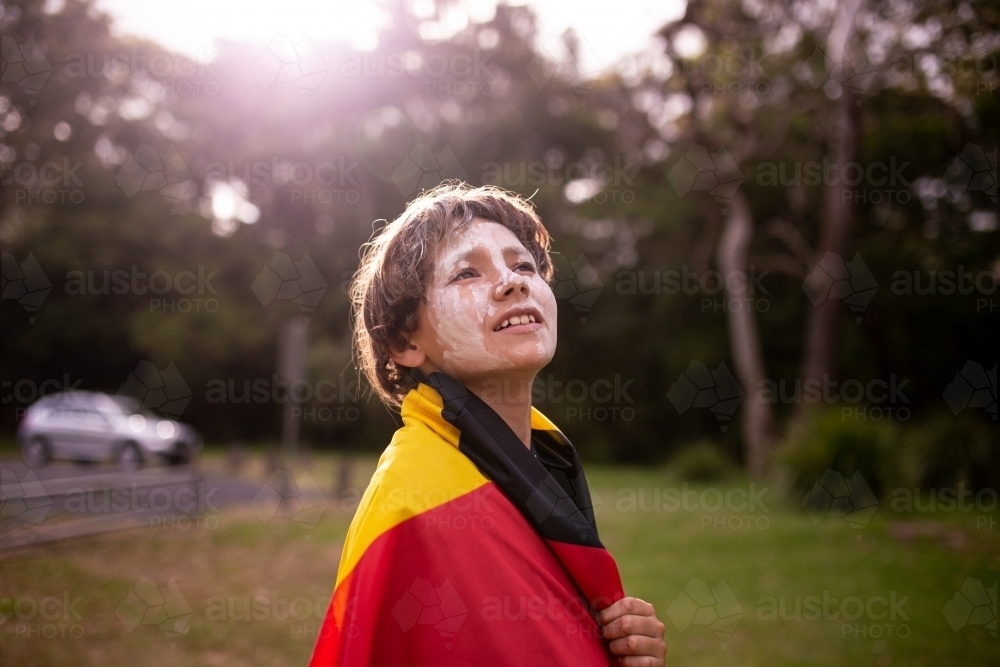 Young Aboriginal boy wearing the Aboriginal flag around his shoulders and looking up - Australian Stock Image