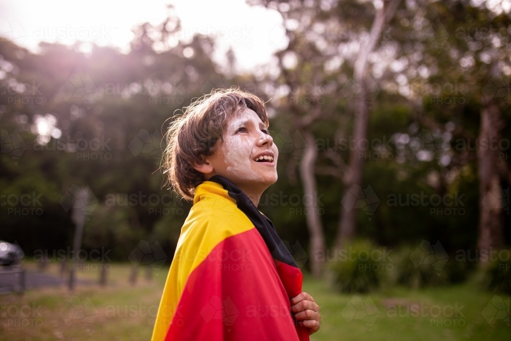 Young Aboriginal boy wearing the Aboriginal flag around his shoulders and smiling - Australian Stock Image