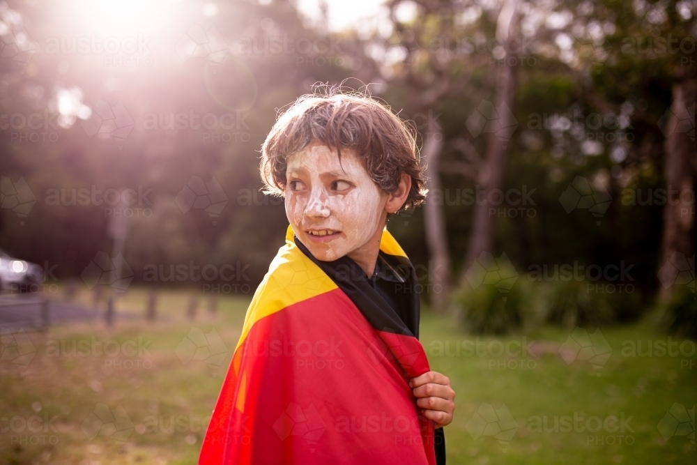 Young Aboriginal boy with white face paint wearing the Aboriginal flag around his shoulders - Australian Stock Image