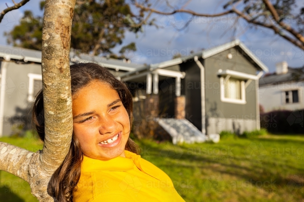 young aboriginal girl in yellow shirt leaning on tree outside her home - Australian Stock Image