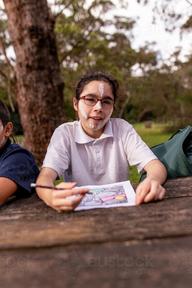 Young Aboriginal girl sitting at an outdoor table drawing and smiling at the camera - Australian Stock Image