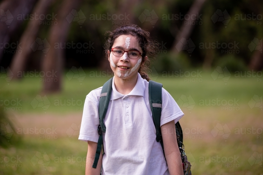 Young Aboriginal girl wearing a backpack, white face paint and glasses smiling at the camera - Australian Stock Image