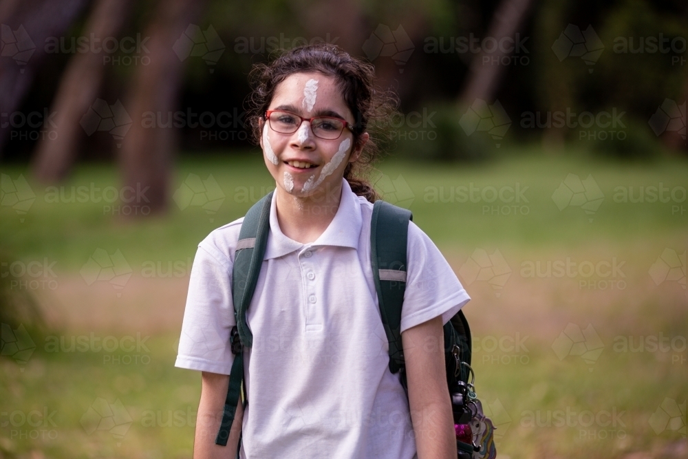 Young Aboriginal girl wearing a backpack, white face paint and glasses smiling at the camera - Australian Stock Image