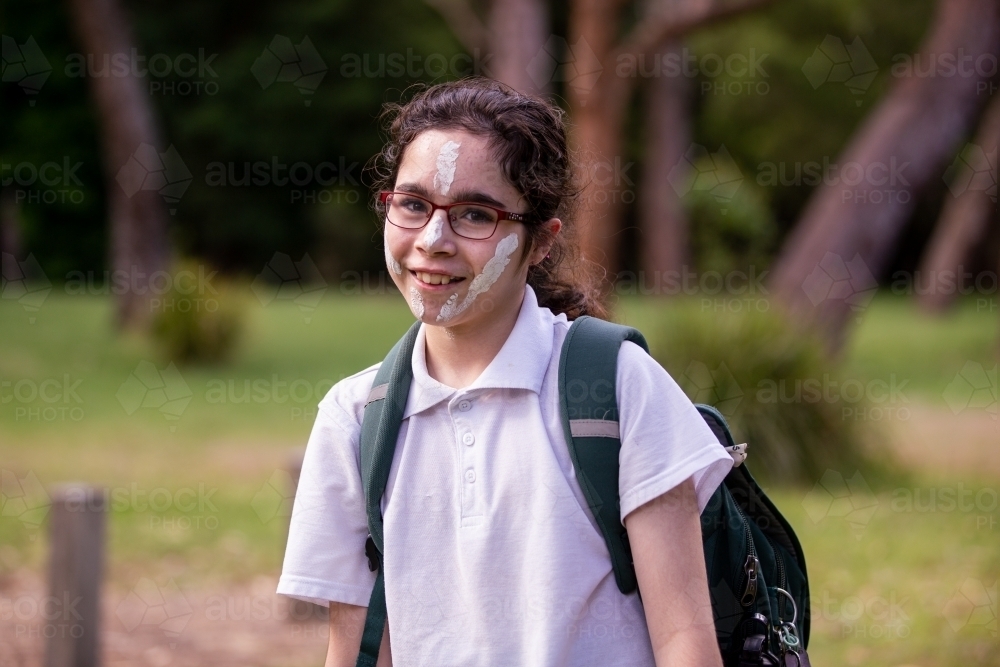 Young Aboriginal girl wearing a backpack, white face paint and glasses smiling at the camera - Australian Stock Image