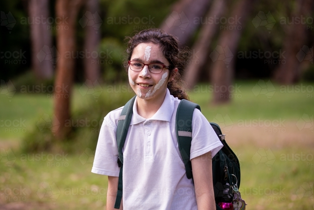 Young Aboriginal girl wearing a backpack, white face paint and glasses smiling at the camera - Australian Stock Image