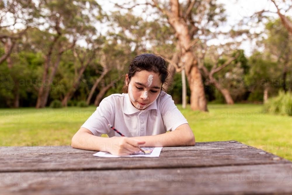 Young Aboriginal girl wearing white face paint drawing at an outdoor table - Australian Stock Image