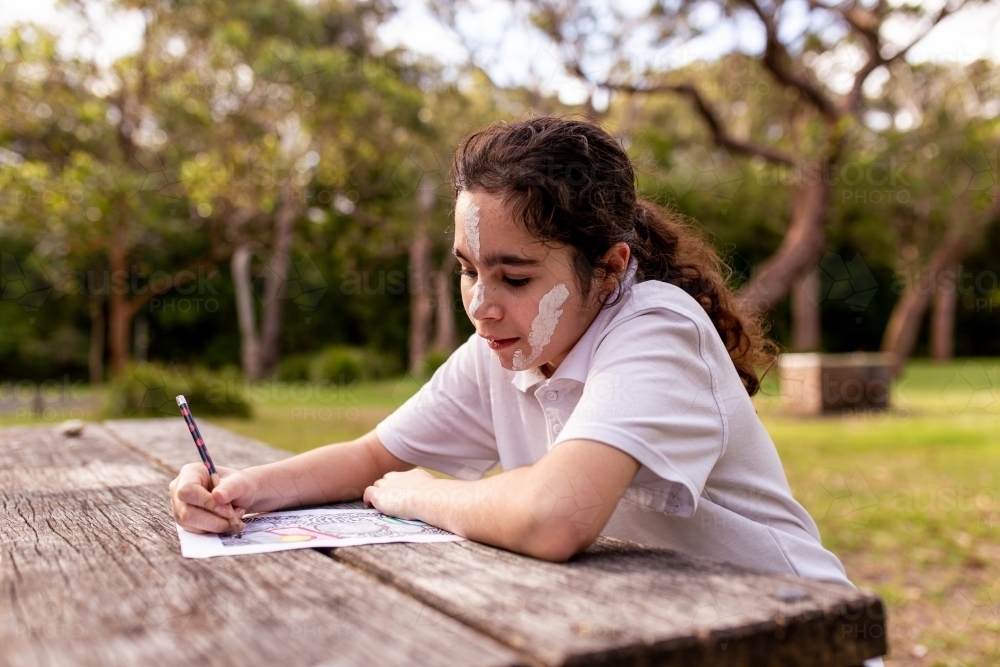 Young Aboriginal girl with face paint drawing at a table outdoors - Australian Stock Image