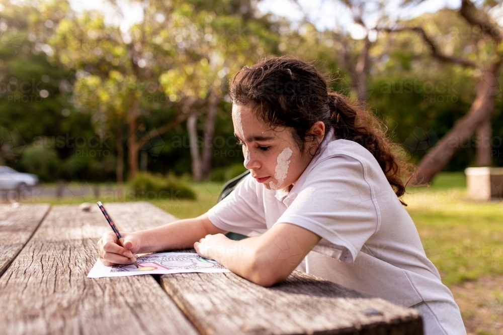 Young Aboriginal girl with white face paint sitting at an outdoor table drawing - Australian Stock Image