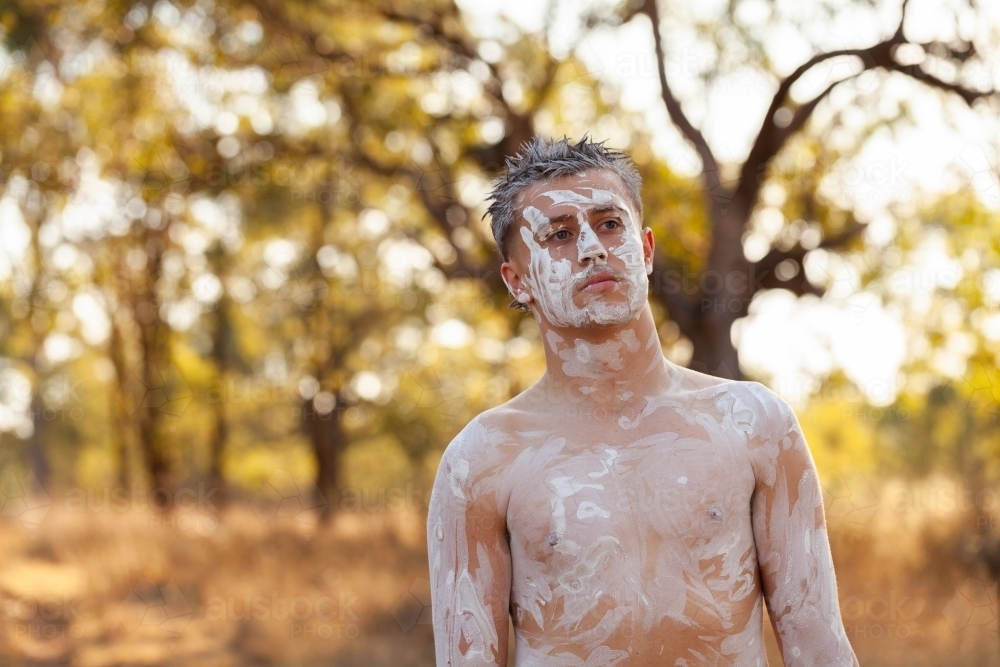 Young aboriginal man with neutral expression standing on country with ochre body paint - Australian Stock Image