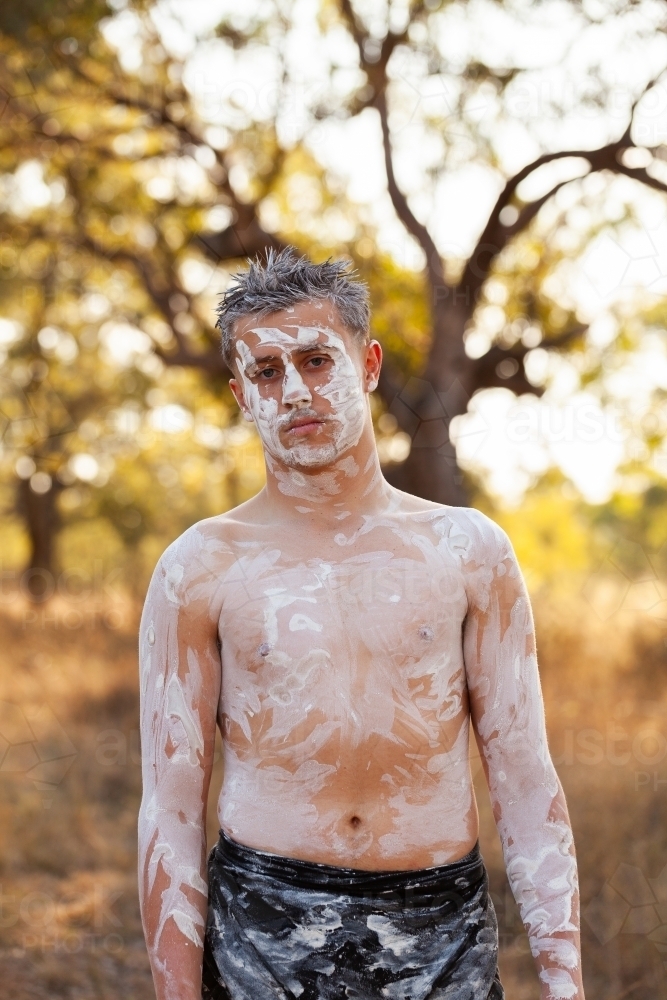 Young aboriginal man with neutral expression standing on country with ochre body paint - Australian Stock Image