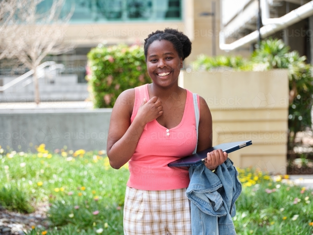 Young African woman holding laptop and jacket in university courtyard - Australian Stock Image