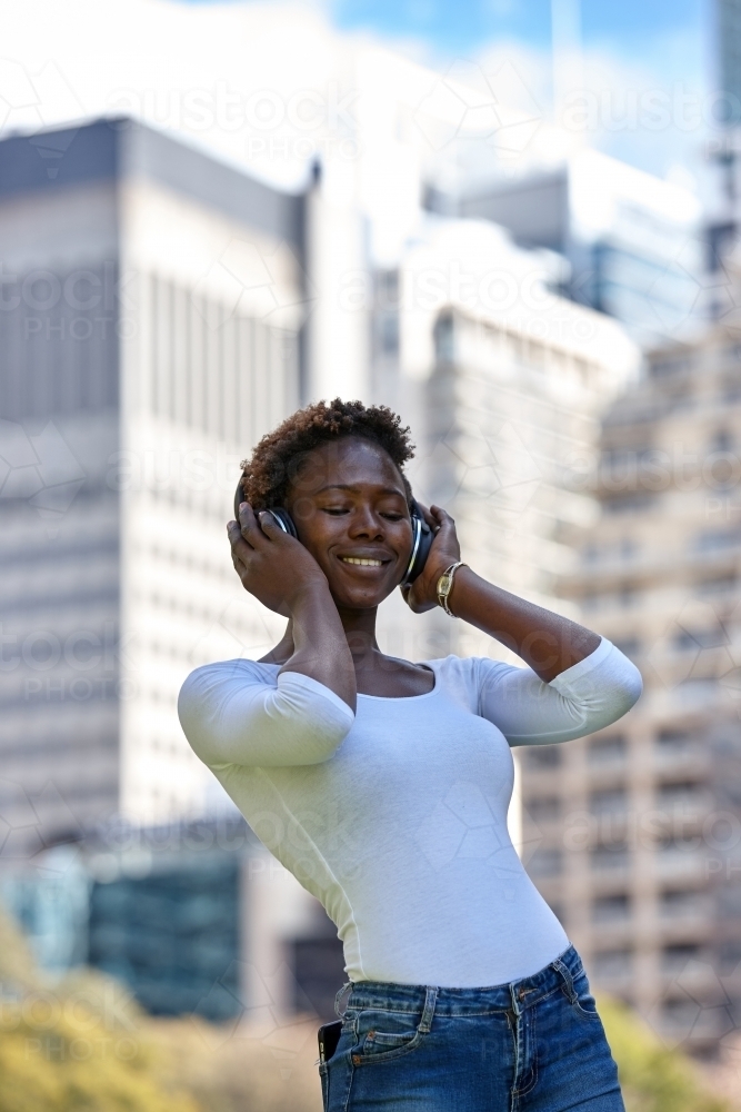 Young African woman listening to music wearing wireless headphones in city - Australian Stock Image