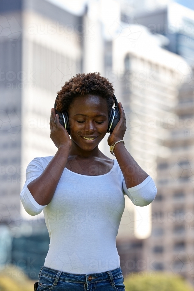 Young African woman listening to music wearing wireless headphones in city - Australian Stock Image