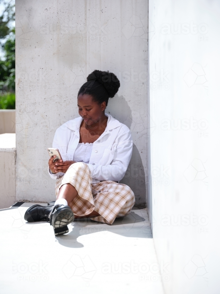 Young African woman using a phone sitting against a concrete wall - Australian Stock Image