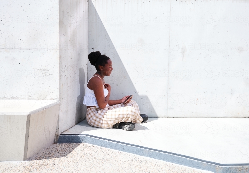 Young African woman using a phone sitting against a concrete wall - Australian Stock Image