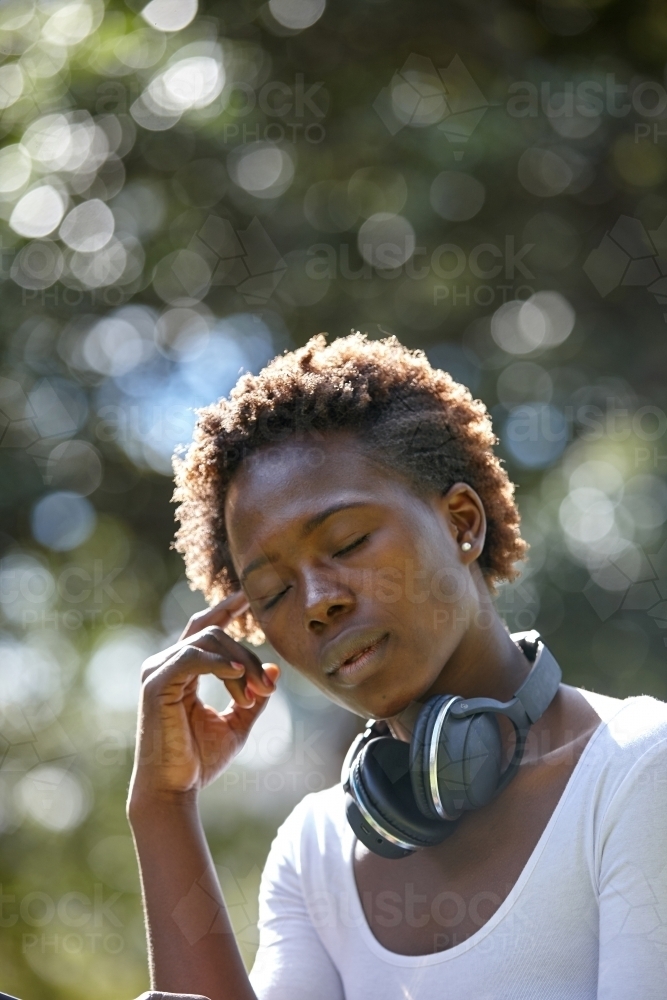 Young African woman with eyes closed wearing wireless headphones at park - Australian Stock Image