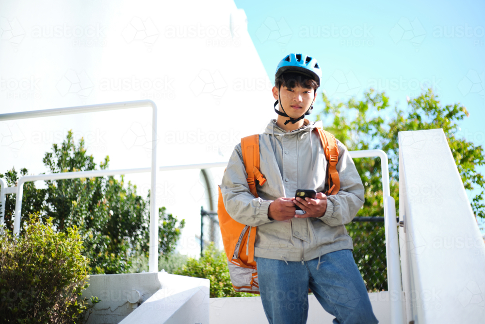 Young Asian delivery guy standing along the stairs while holding his phone - Australian Stock Image
