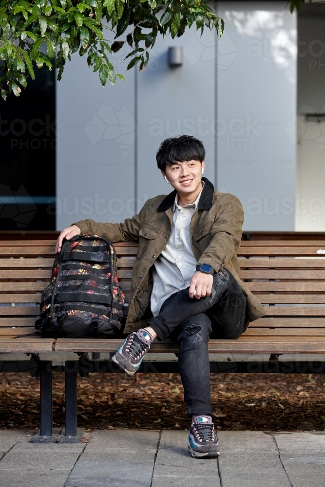 Young Asian man smiling sitting on park bench whilst listening to wireless headphones - Australian Stock Image