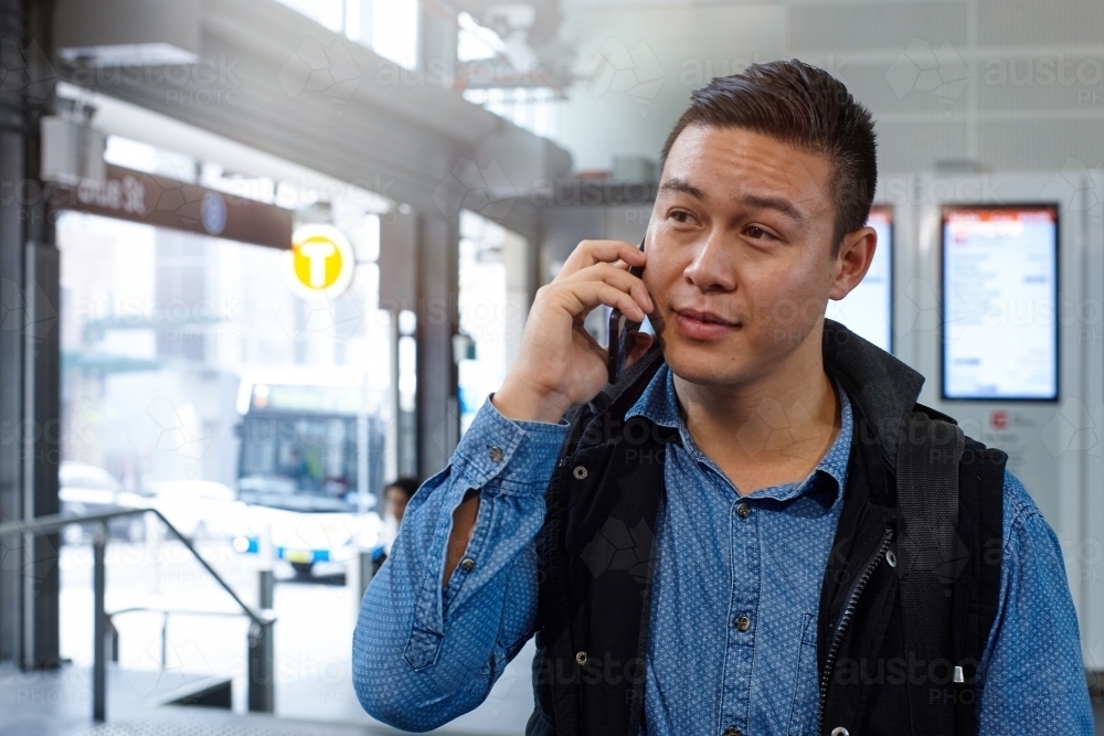 Young Asian man talking on mobile phone at train station entrance - Australian Stock Image