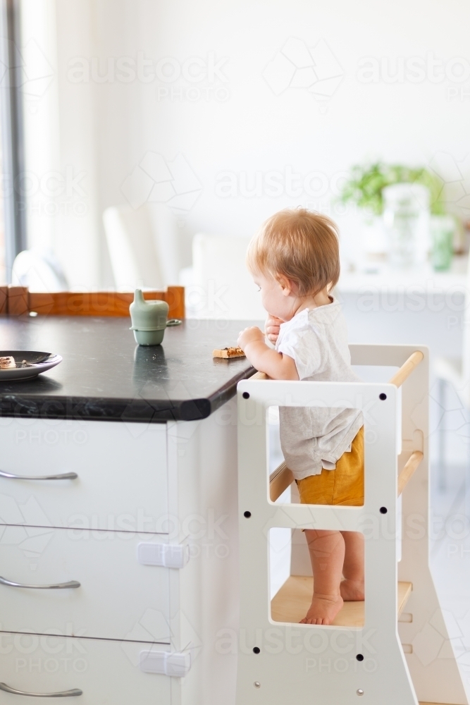 Young australian baby eating vegemite toast at kitchen bench standing on Montessori learning tower - Australian Stock Image