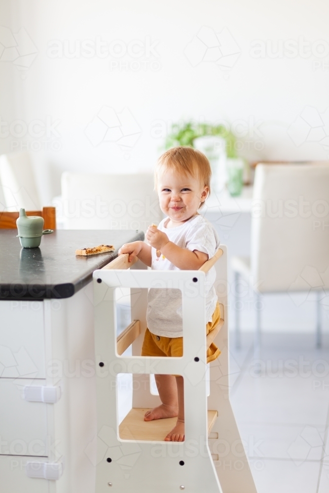 Young australian baby eating vegemite toast at kitchen bench standing on Montessori learning tower - Australian Stock Image