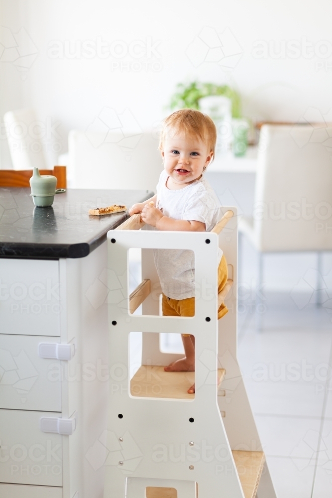 Young australian baby eating vegemite toast at kitchen bench standing on Montessori learning tower - Australian Stock Image