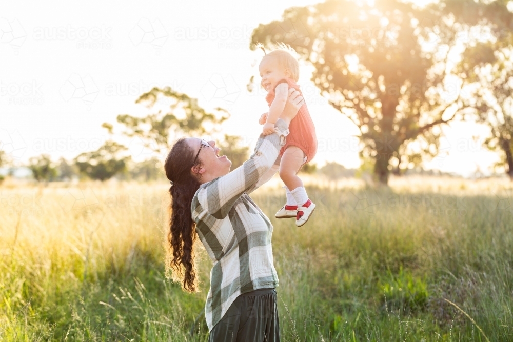 Young australian mother on farm lifting up her one year old child in sunlit paddock in autumn - Australian Stock Image