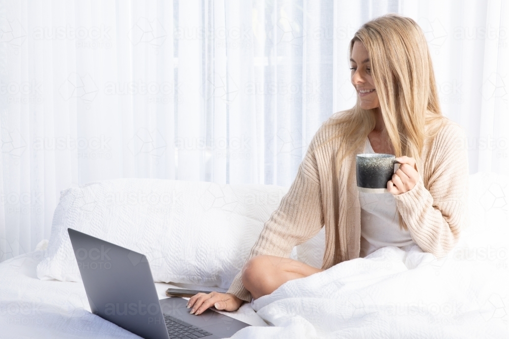 Young blonde lady typing on a laptop in white bed - Australian Stock Image