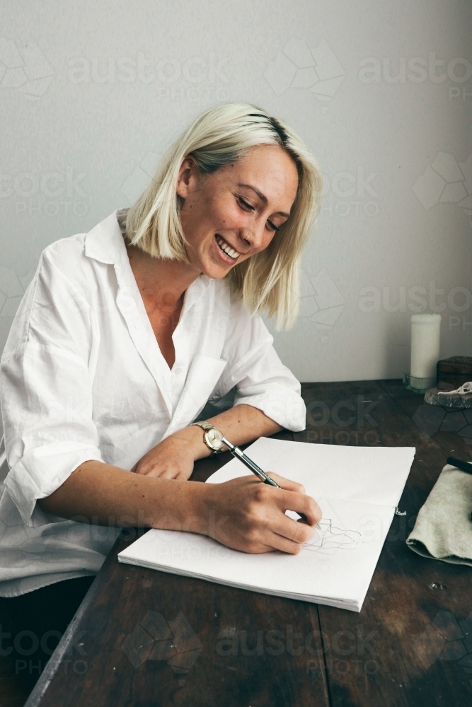 Young blonde woman laughing as she sits at a desk drawing in a sketch book - Australian Stock Image