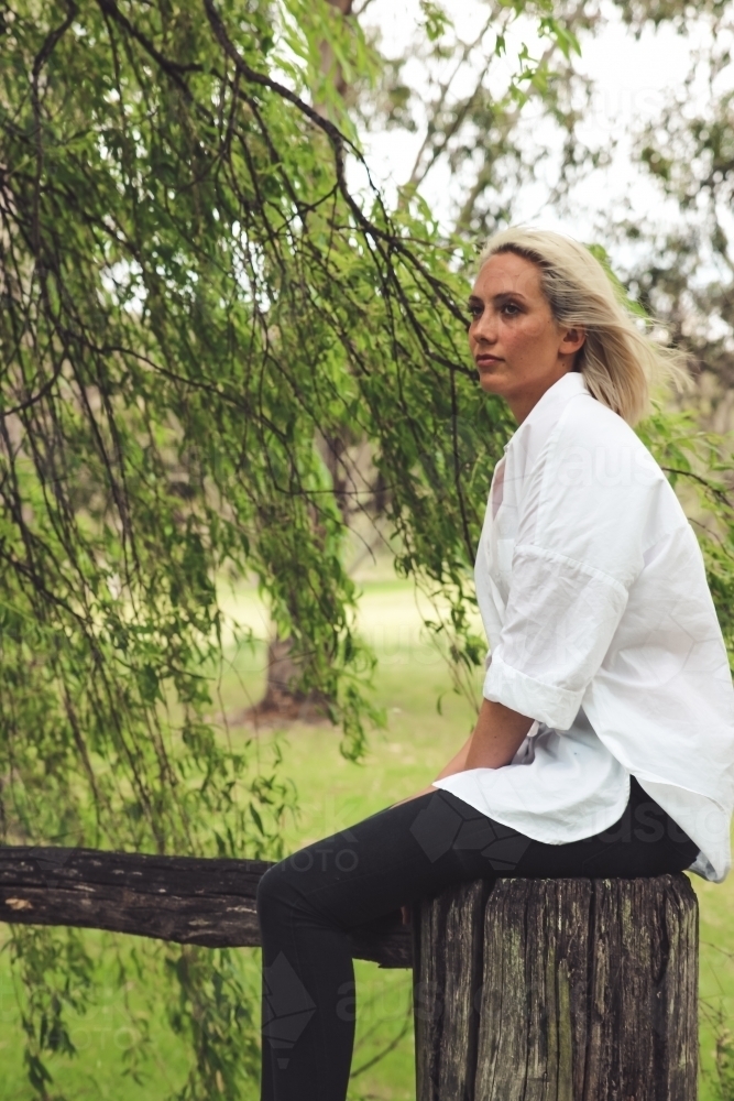 Young blonde woman looking away, sitting on a fence post as wind blows her hair - Australian Stock Image