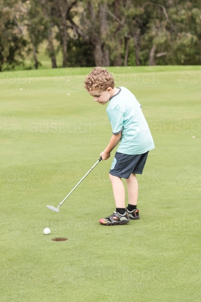 Young boy holding golf club putter hitting ball into hole on green - Australian Stock Image