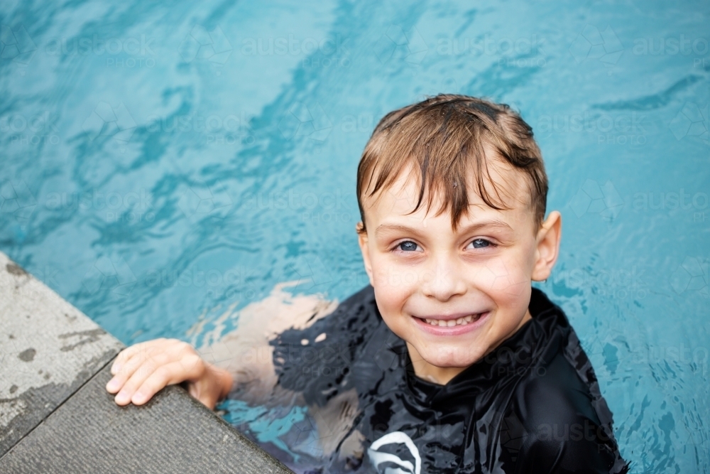 Young boy in a swimming pool, looking and smiling at the camera - Australian Stock Image